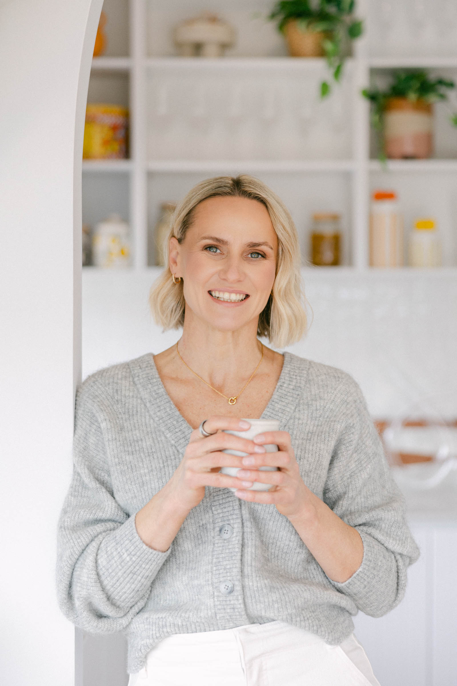 Zelda Green, financial educator for photographers and money mindset coach, smiling at camera in a brightly lit home wearing a grey cardigan and white jeans